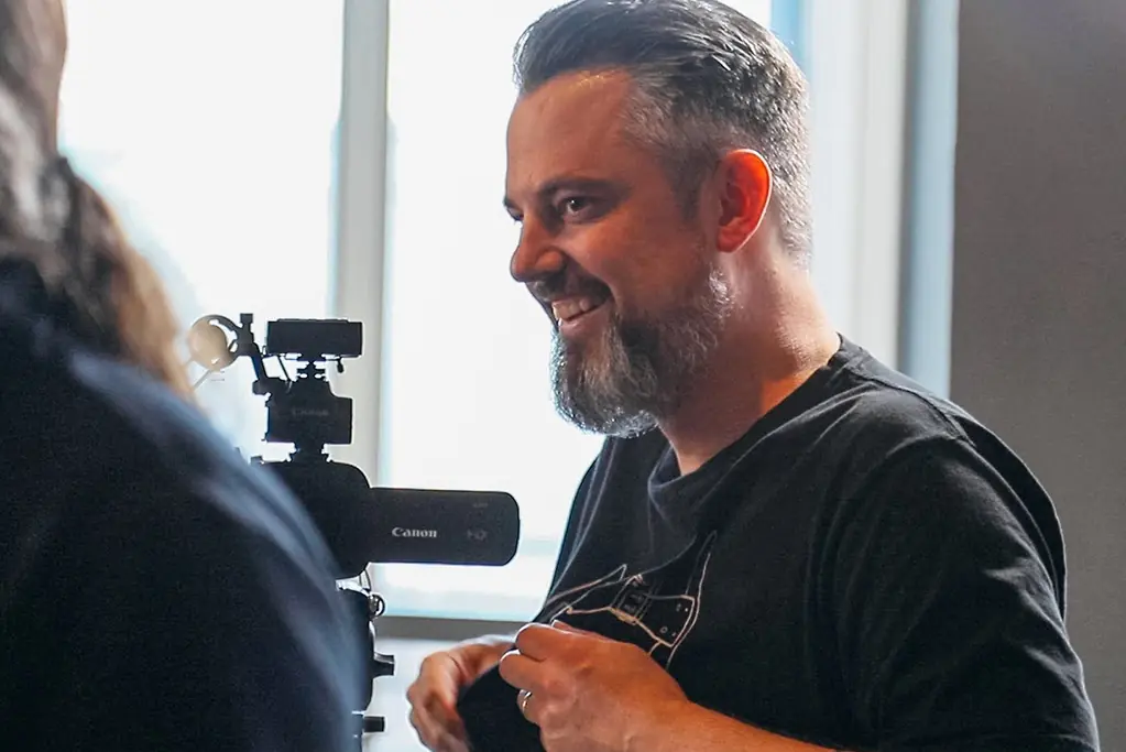 Man smiling and talking beside a colleague, with a small Canon camera on a tripod set up in front of a bright window