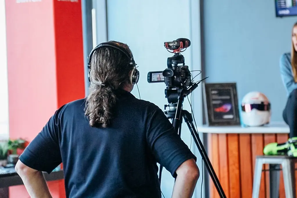 Camera operator wearing a headset filming a woman being interviewed indoors, with a racing helmet and framed award displayed nearby