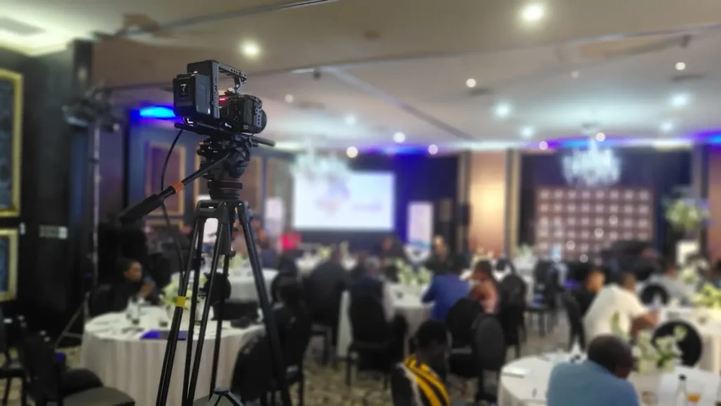 Camera on a tripod set up in an event ballroom, with guests seated at tables and a presentation screen in the background.